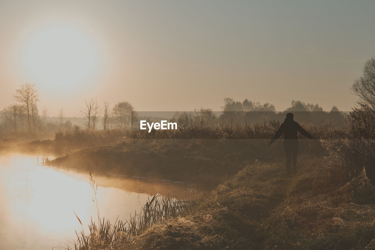 Rear view of silhouette man standing by lake against sky at sunrise