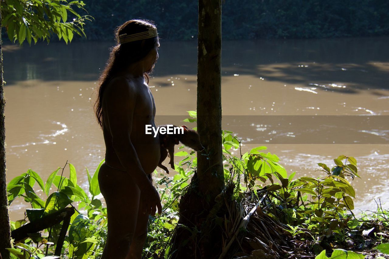 Side view of shirtless man holding dog while standing by lake