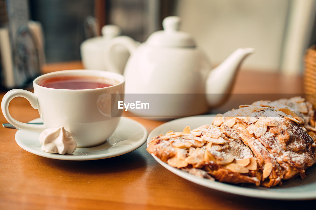 White cup with hot tea, tea pot and fresh croissant on plate in cafe in the morning, close up