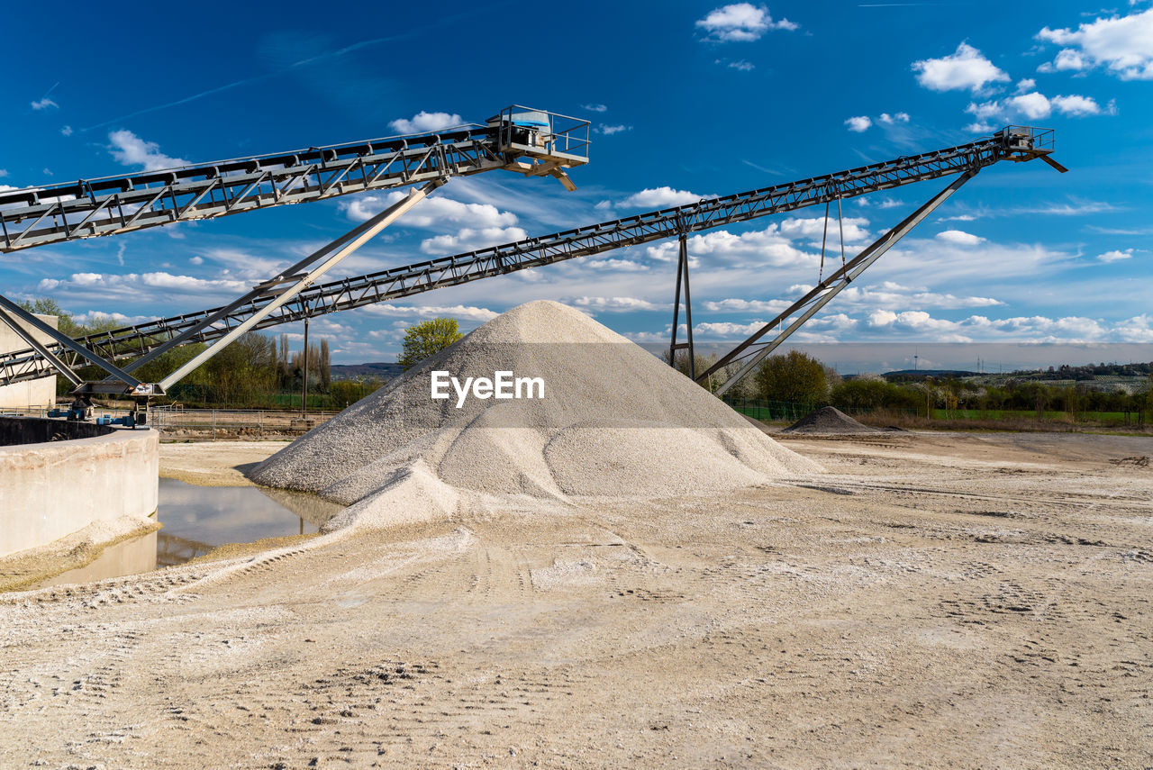 Conveyor over heaps of gravel on blue sky at an industrial cement plant.