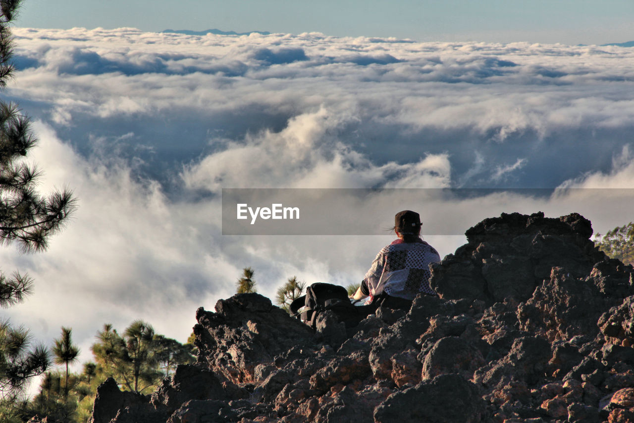 Rear view of person sitting on mountain against cloudscape