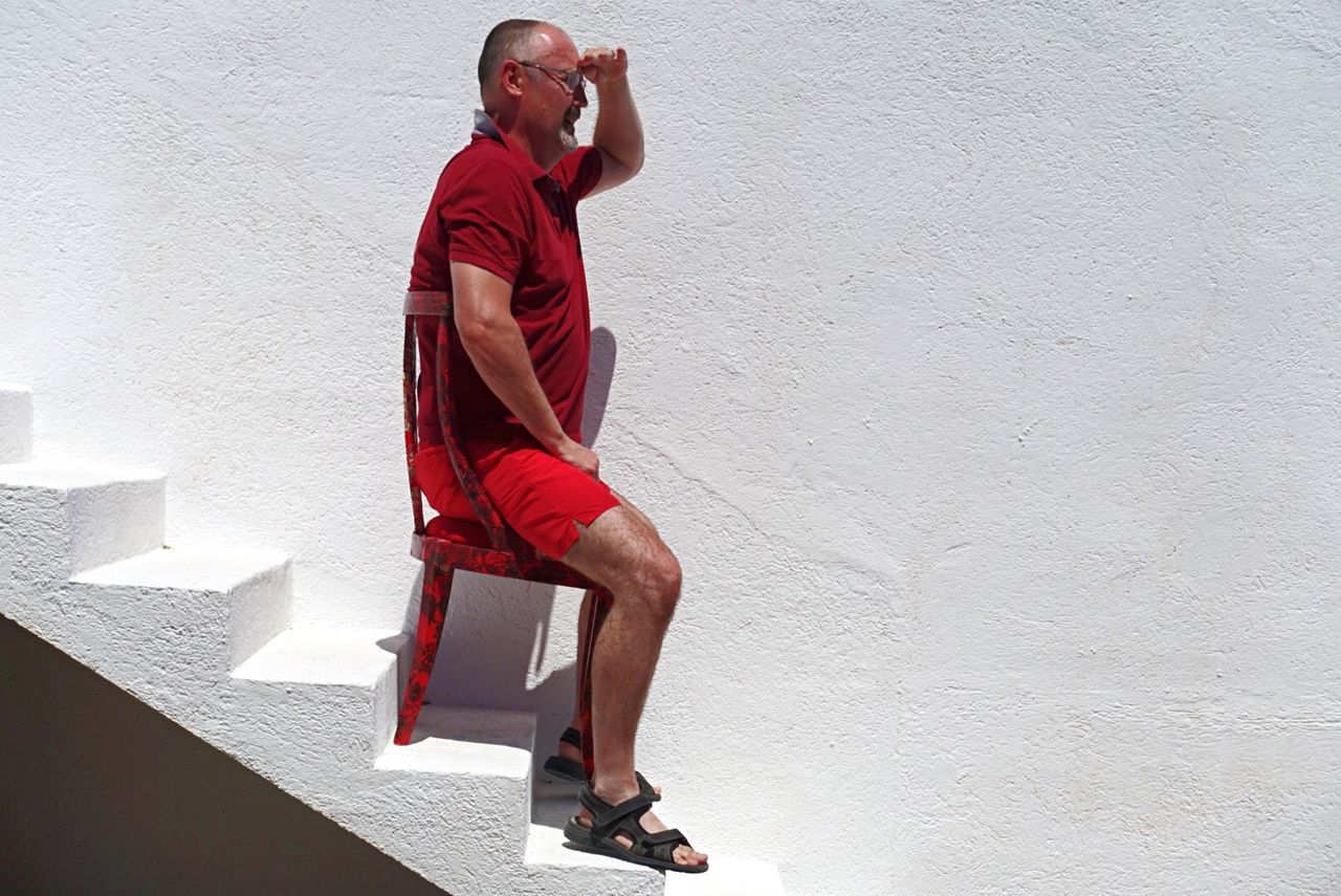 Side view full length of man shielding eyes while sitting on chair at steps against wall