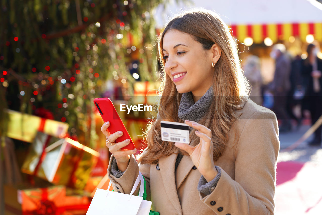 Beautiful smiling girl paying her gifts with mobile phone with christmas markets in the background.