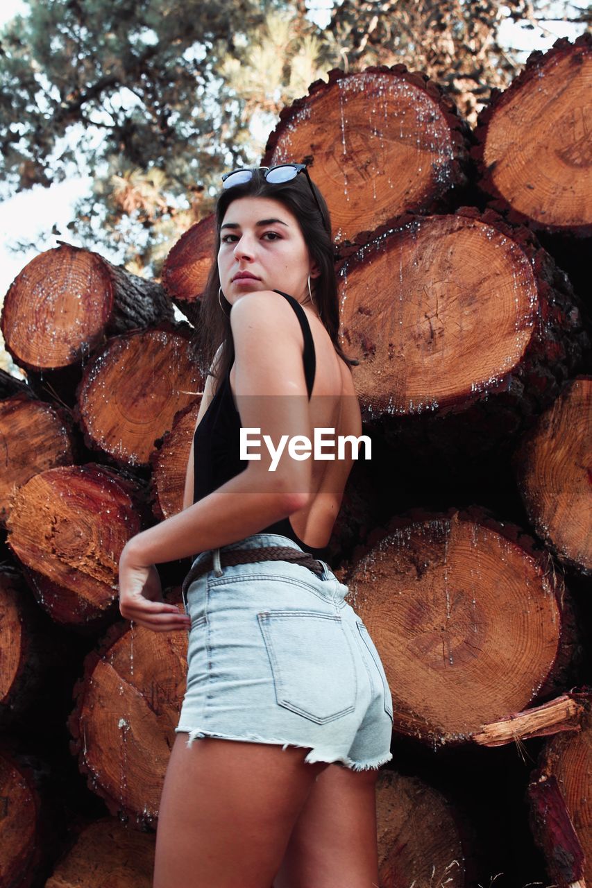 Low angle portrait of young woman standing by logs in forest