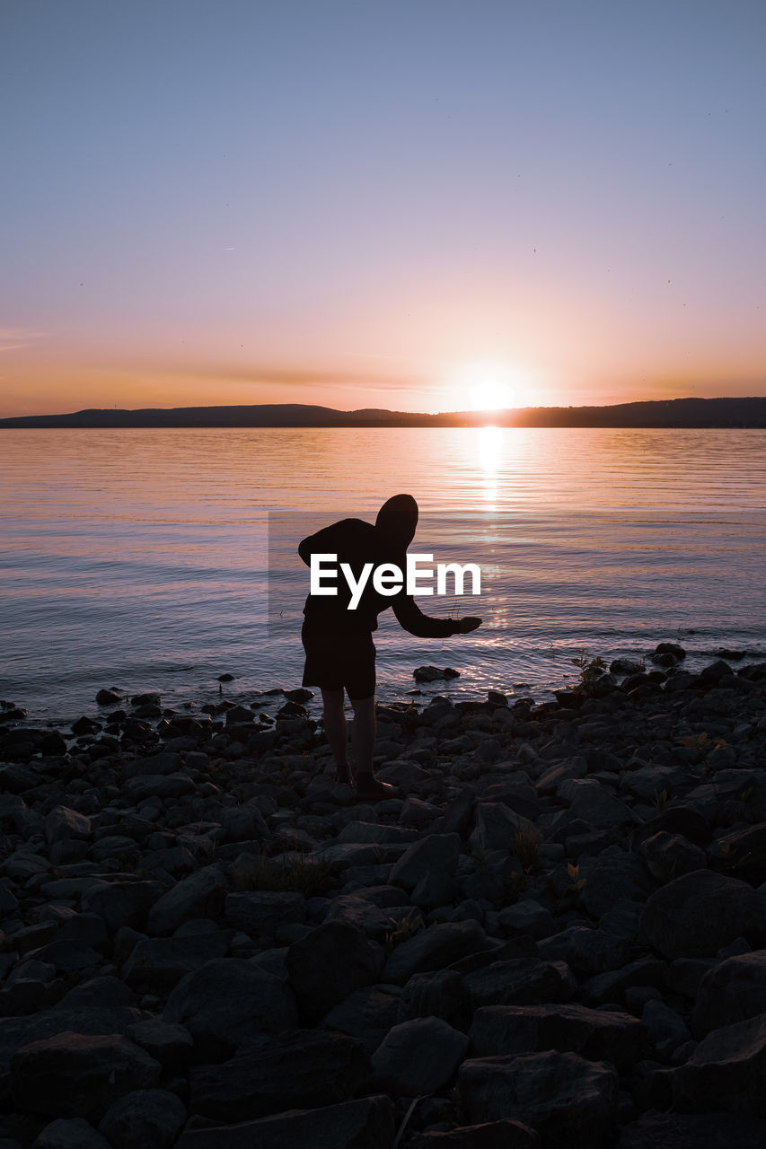 Man standing on rock at lakeshore against sky during sunset