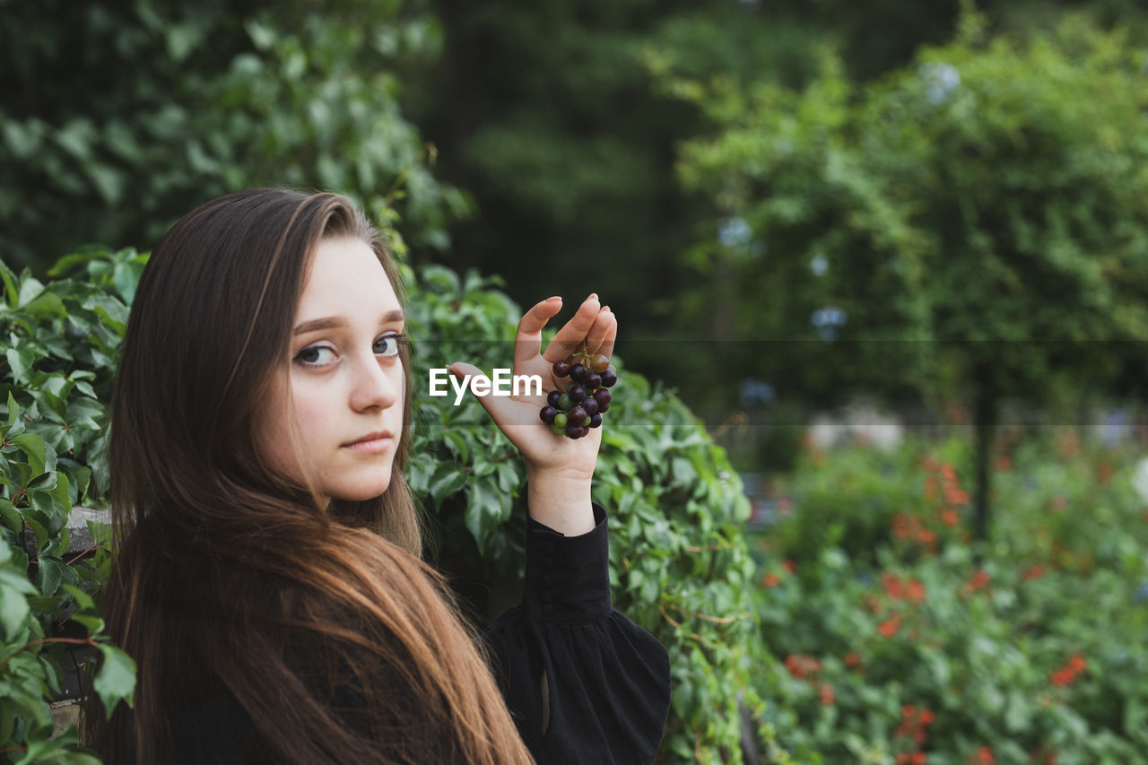 Portrait of smiling young woman holding fruits standing against plants