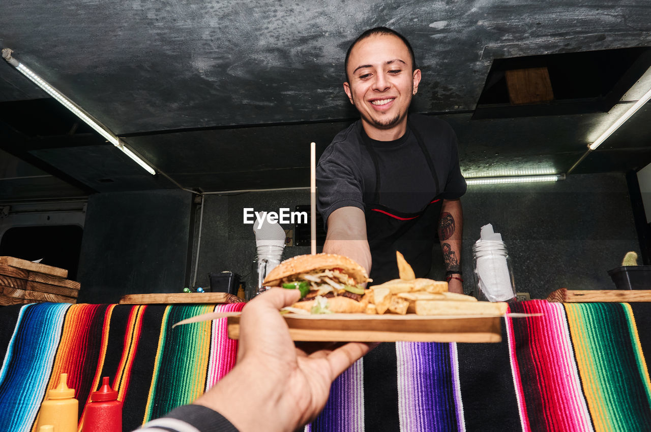 Hand of faceless unrecognizable buyer getting order with fast food from happy ethnic salesman at truck counter