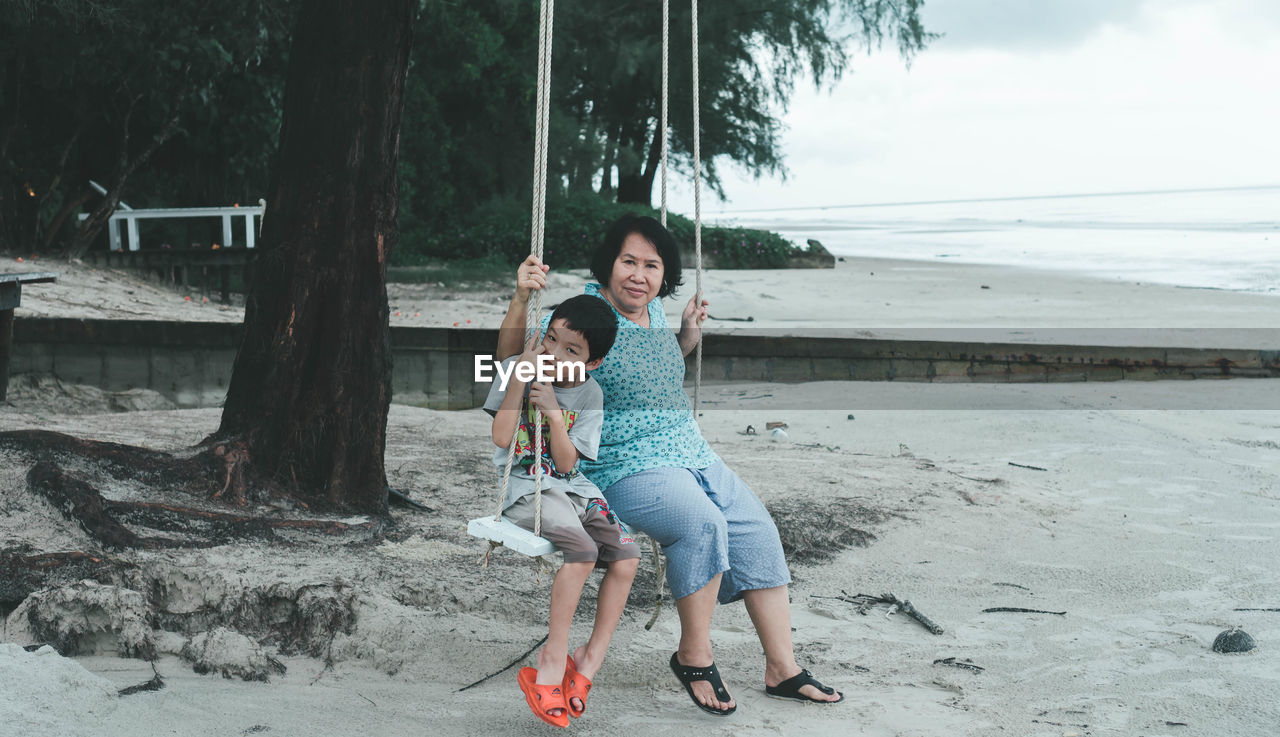 Full length portrait of woman sitting with grandson on swing at beach