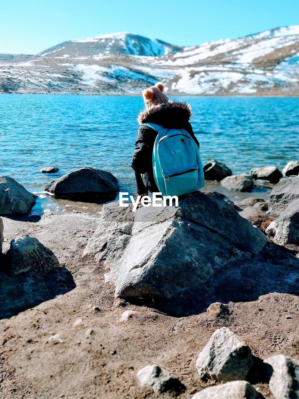 Rear view of teenage girl sitting at lakeshore against snowcapped mountains
