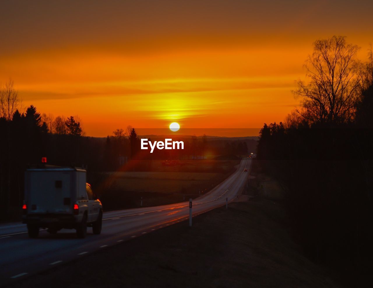 Pick-up truck on country road against orange sky during sunset