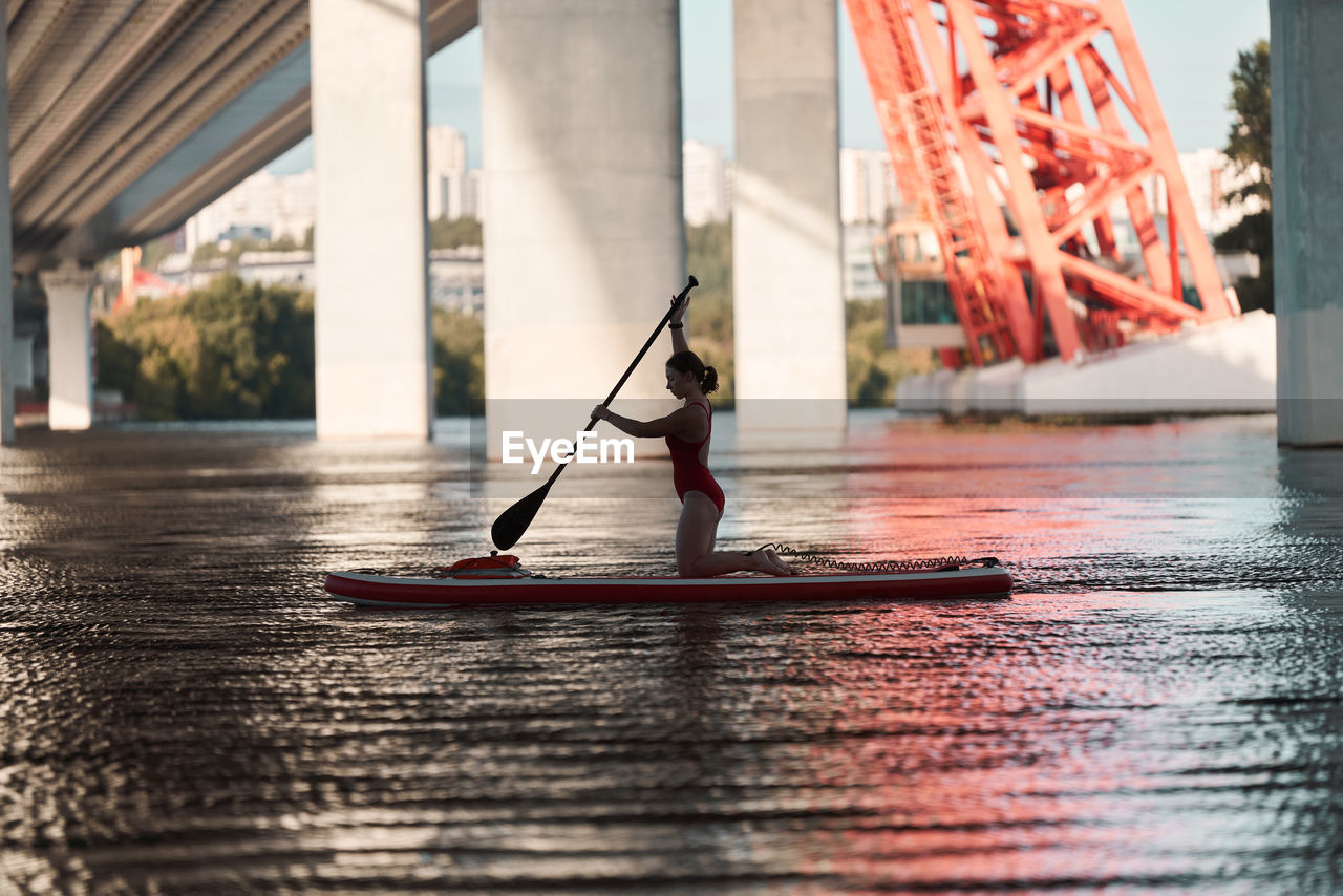 Side view of woman standing on paddle board in river and rowing with paddle under bridge in city