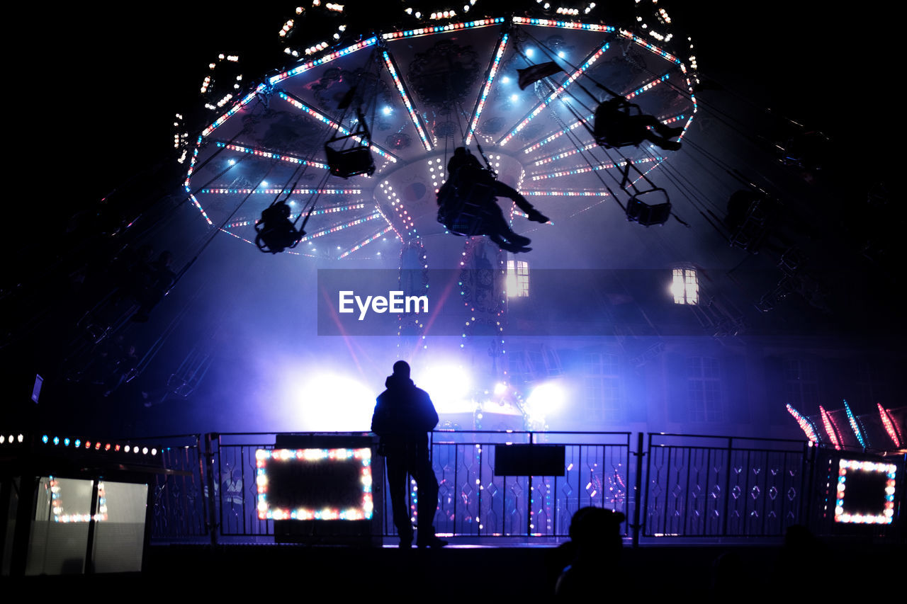 Silhouette man standing against illuminated chain swing ride at night