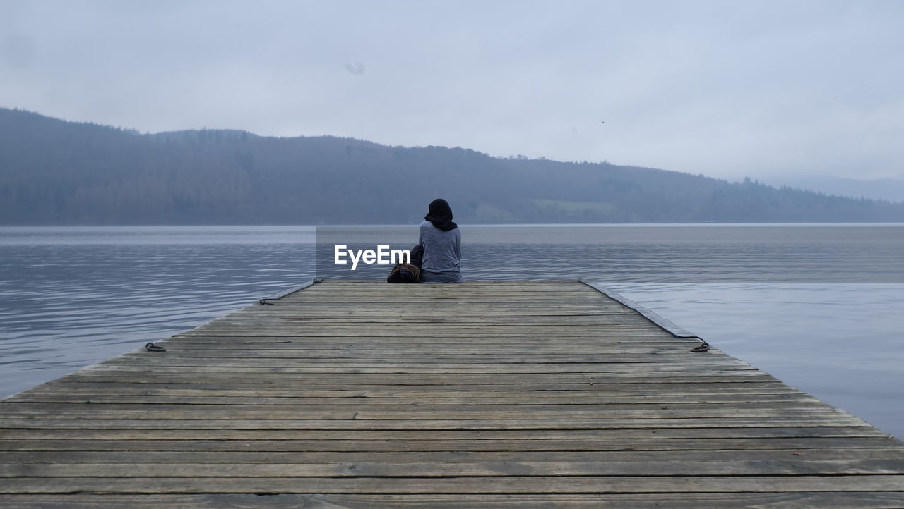 Rear view of woman sitting on pier over lake during foggy weather