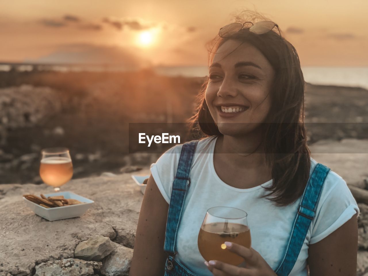 Smiling young woman holding drink against sky at sunset