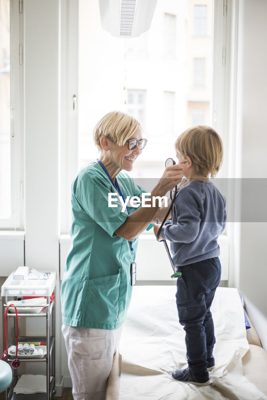 Smiling female doctor holding stethoscope to boy's ear while standing in hospital