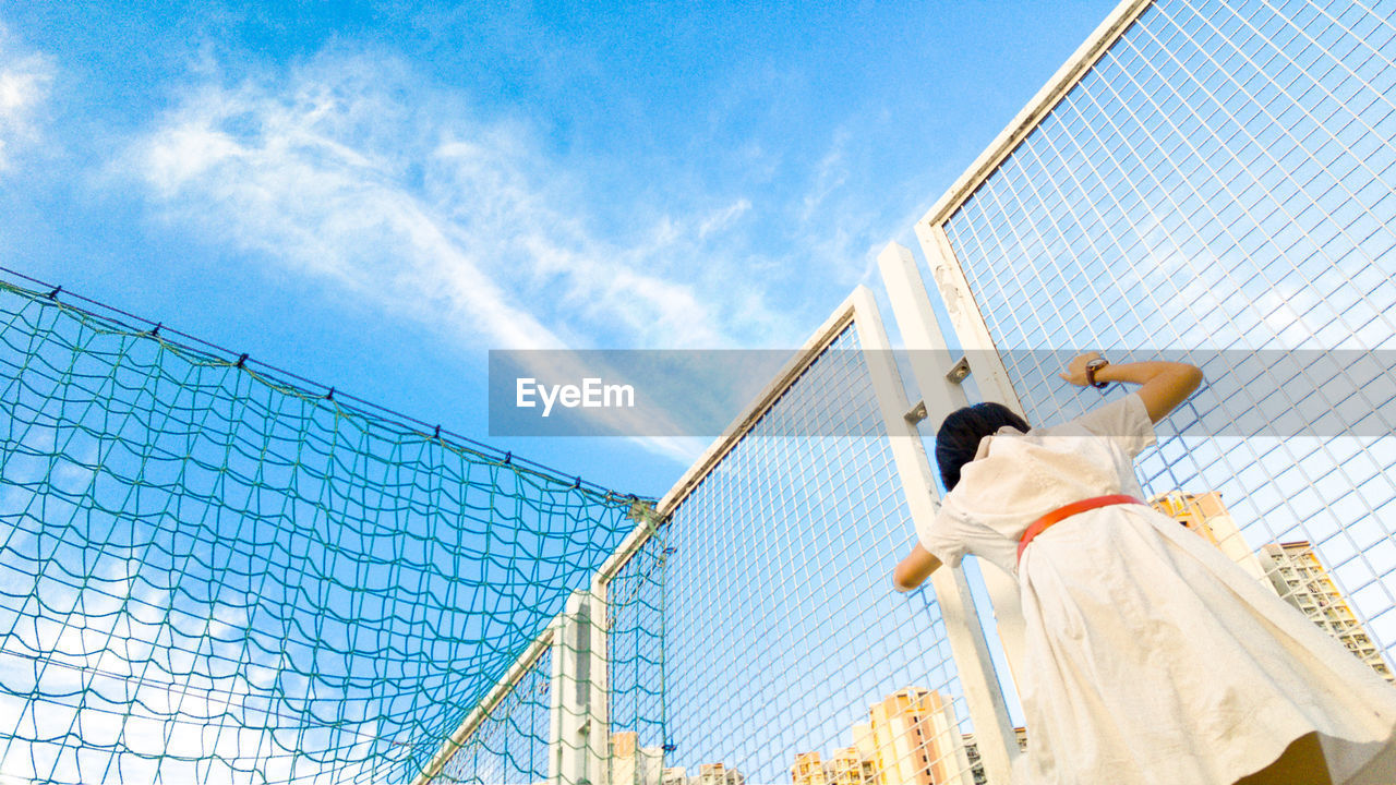 Low angle view of girl standing by fence at schoolyard against sky