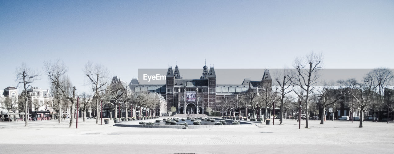 BARE TREES ON SNOW COVERED FIELD AGAINST BUILDINGS