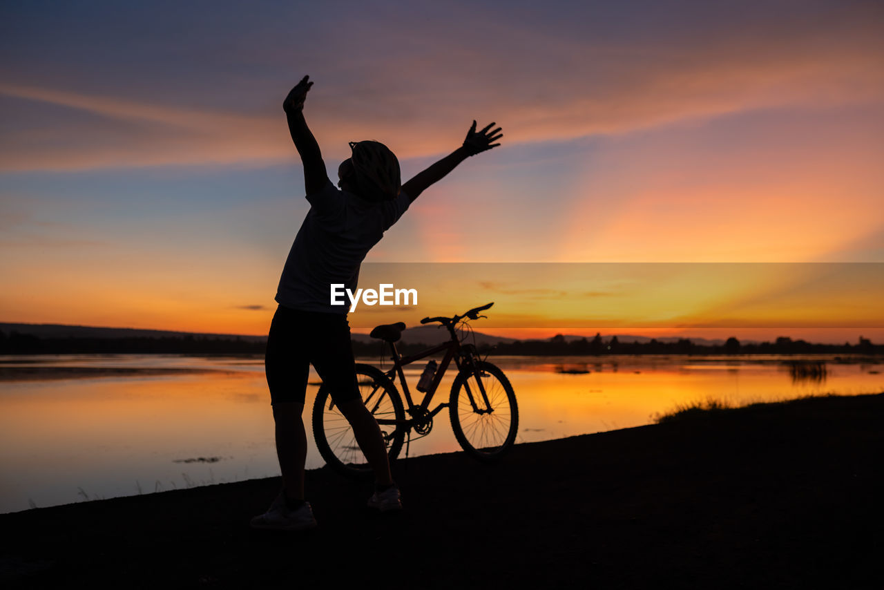 Rear view of man with arms raised standing by bicycle at lakeshore