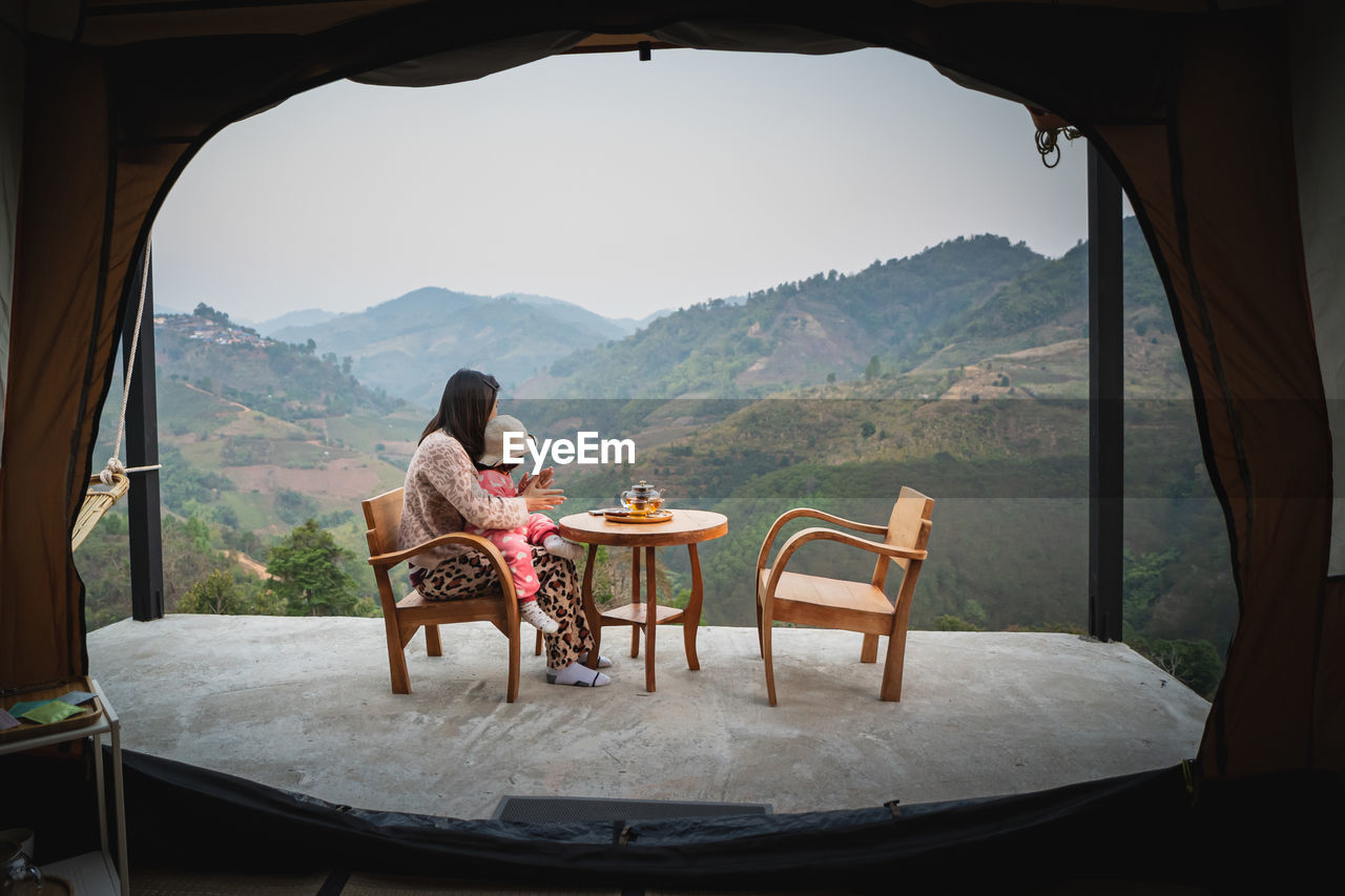 REAR VIEW OF WOMAN SITTING ON CHAIR IN MOUNTAINS
