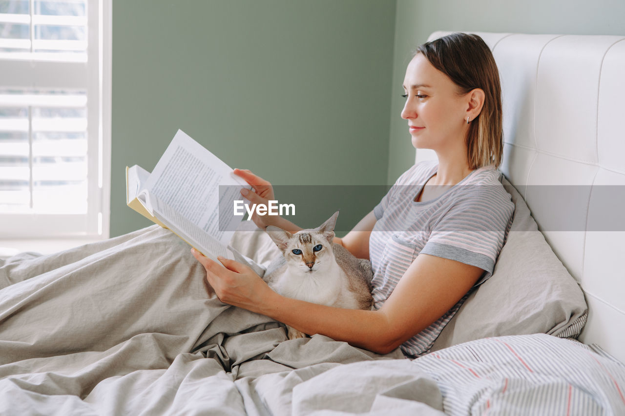 Woman reading book while sitting on bed with cat
