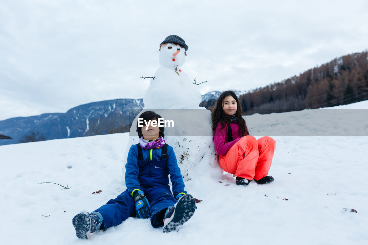 Two children sitting next to snowman they made on top of snowy mountain
