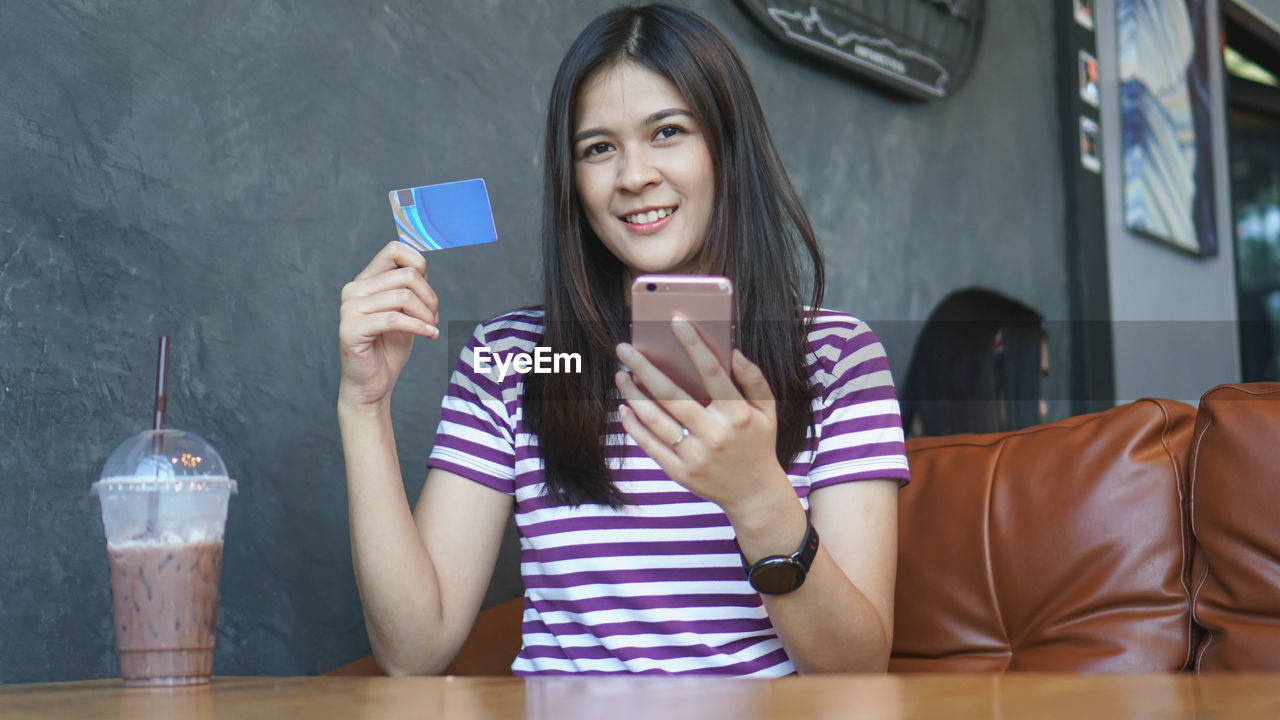 Portrait of smiling young woman holding credit card while using smart phone at table in cafe