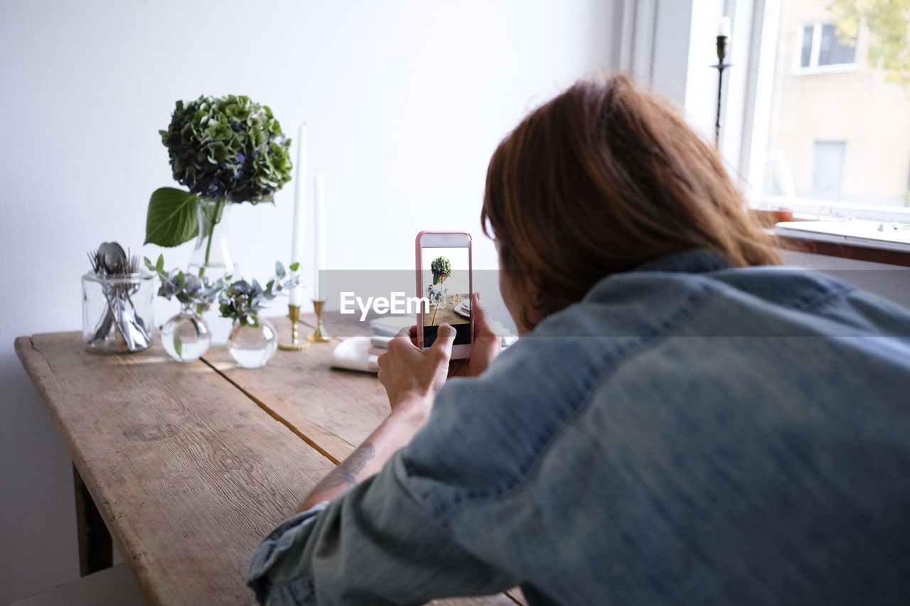 Rear view of blogger photographing flower vases on table through mobile phone