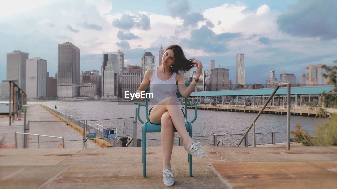 Portrait of young woman sitting on chair at harbor with cityscape in background against sky