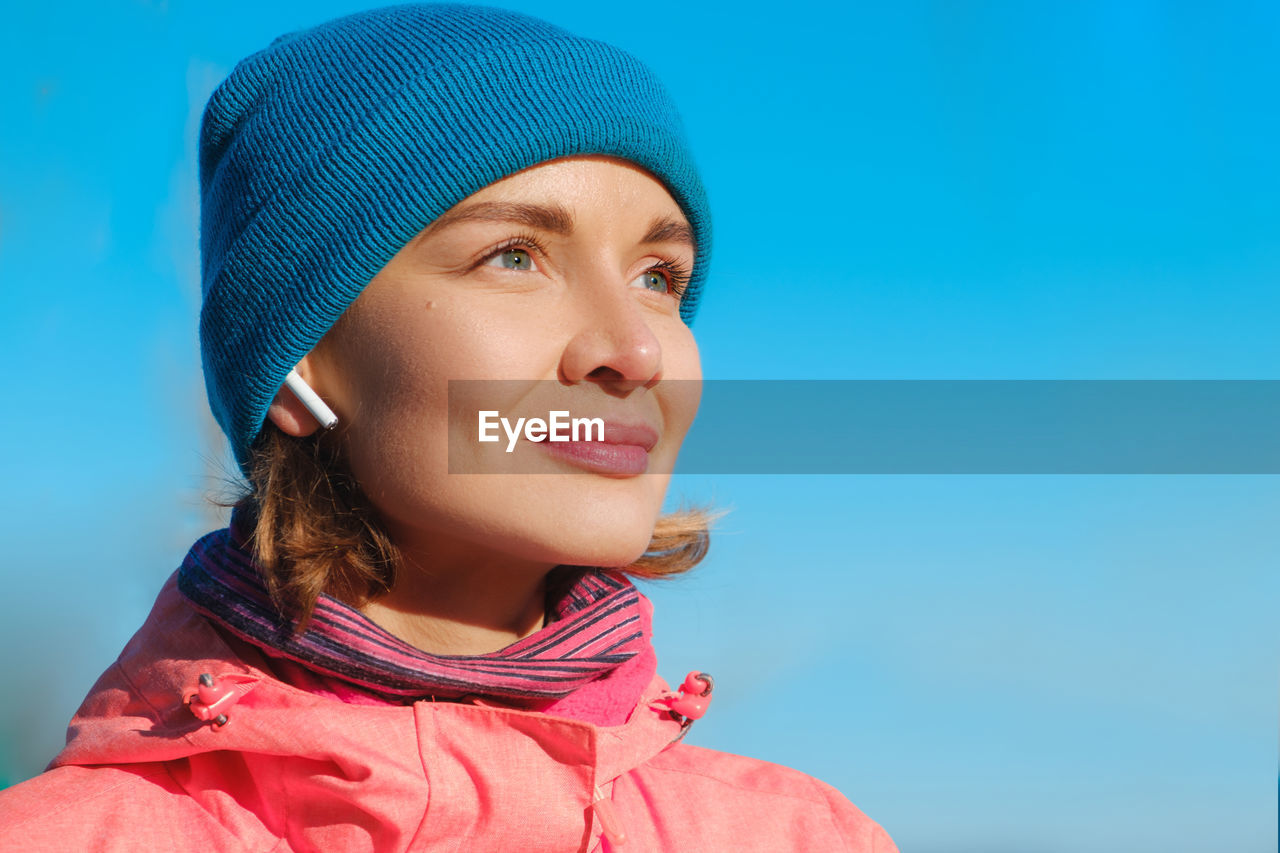 Close-up of smiling woman listening music against blue sky