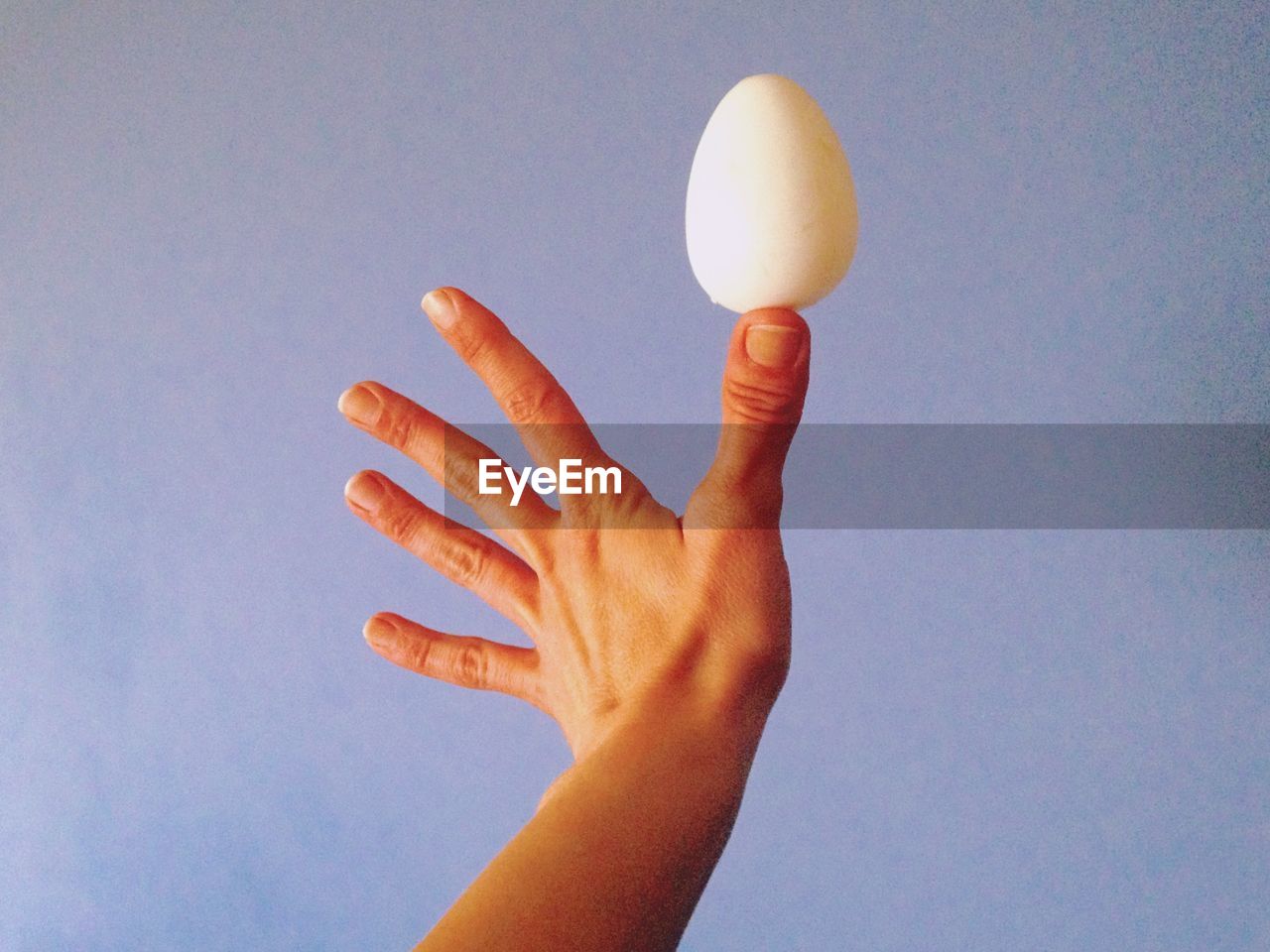 Cropped image of hand balancing egg on thumb against blue background