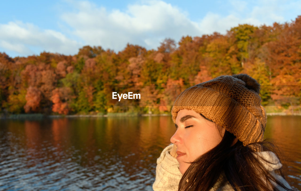 Outdoor portrait of beautiful young woman enjoying sunlight by lake in autumn