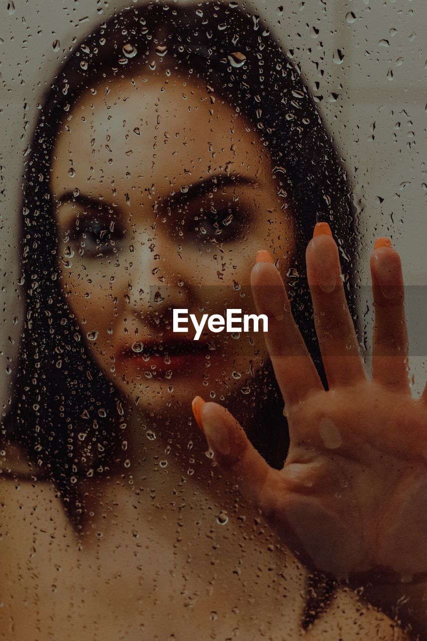 Close-up portrait of young woman seen through wet glass