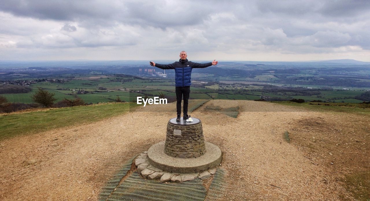 Man with arms outstretched standing on built structure at field against sky