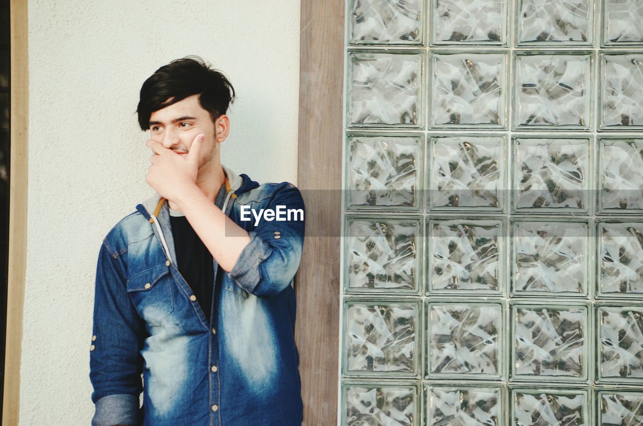 Young man covering mouth looking away while standing against wall