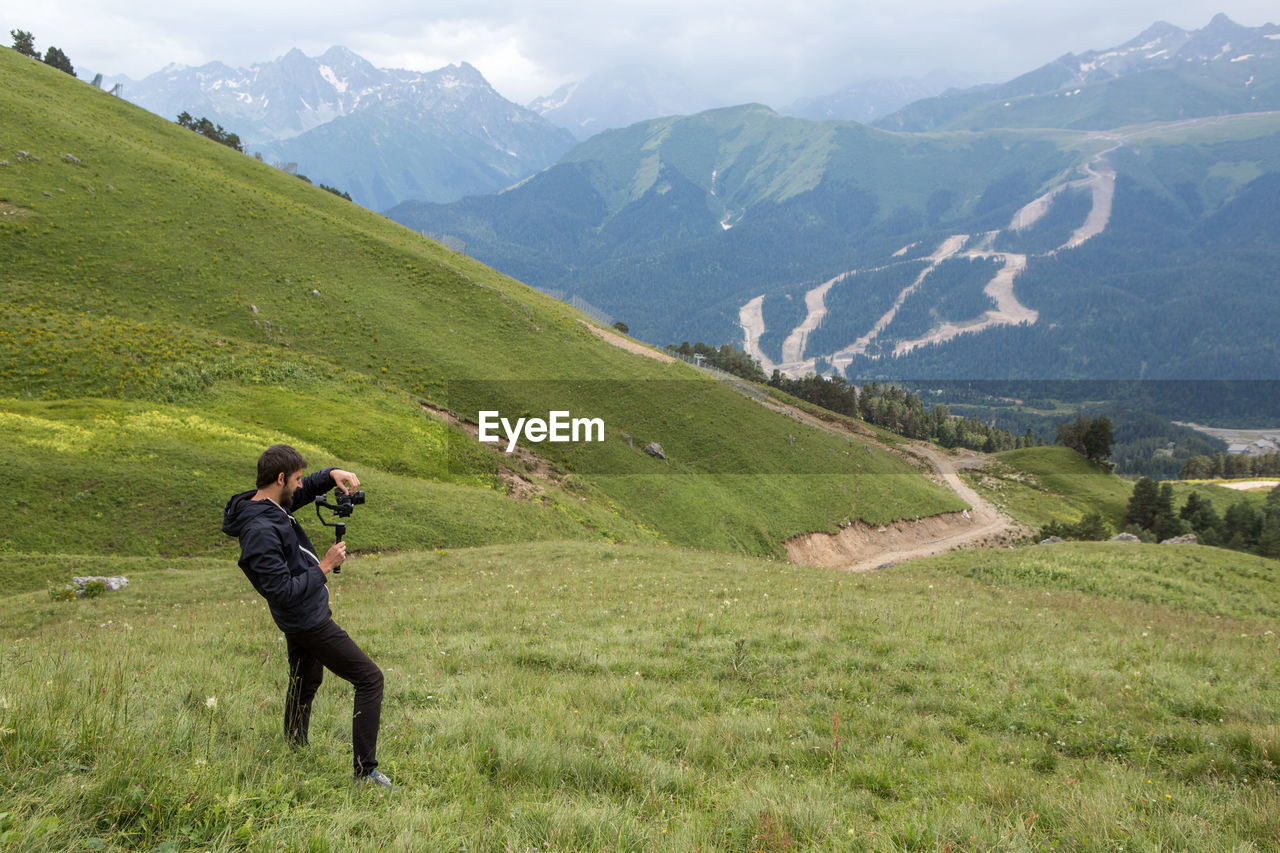 Guy kameraman in a jacket stands in the mountains on a green meadow and shoots a video on steadicam