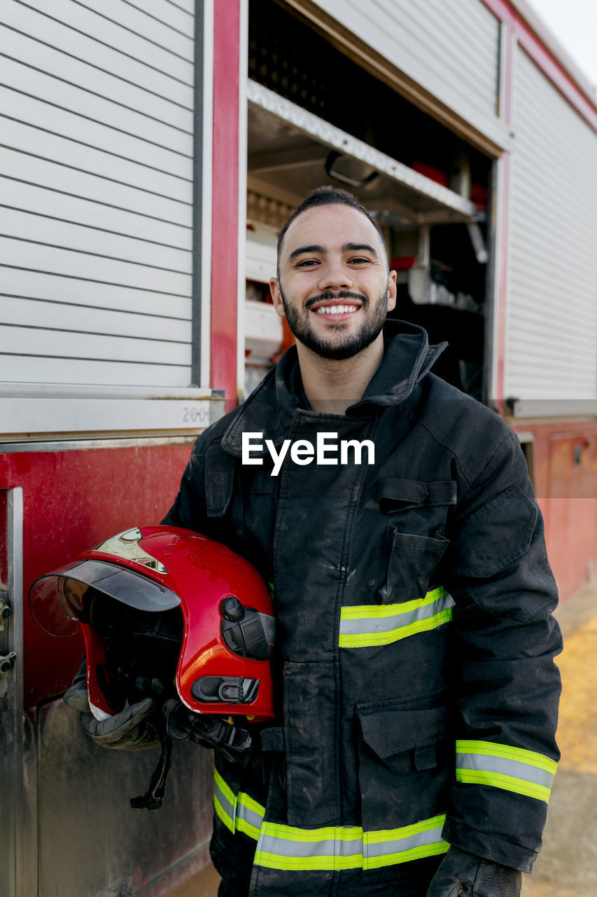 Cheerful bearded young firefighter standing near fire truck and holding red hardhat in hand while smiling and looking at camera