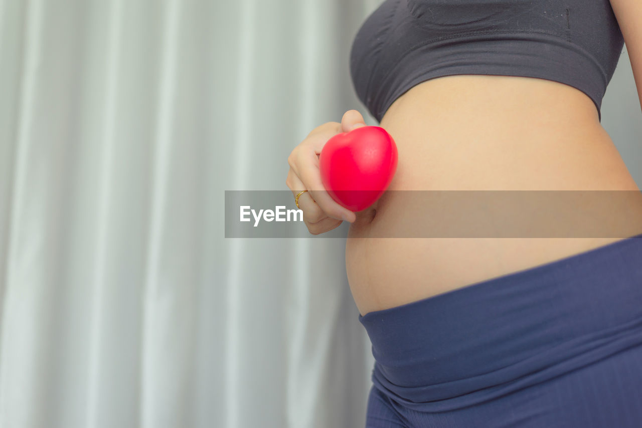 Midsection of pregnant woman holding heart shape against abdomen while standing at home