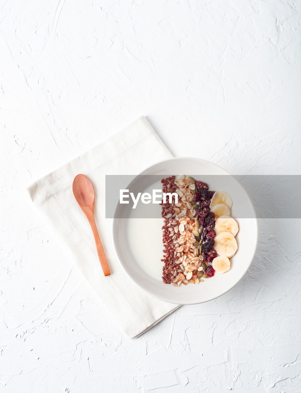 Top view of bowl with white yogurt topped with assorted cereals and sliced banana served for healthy breakfast on table with wooden spoon