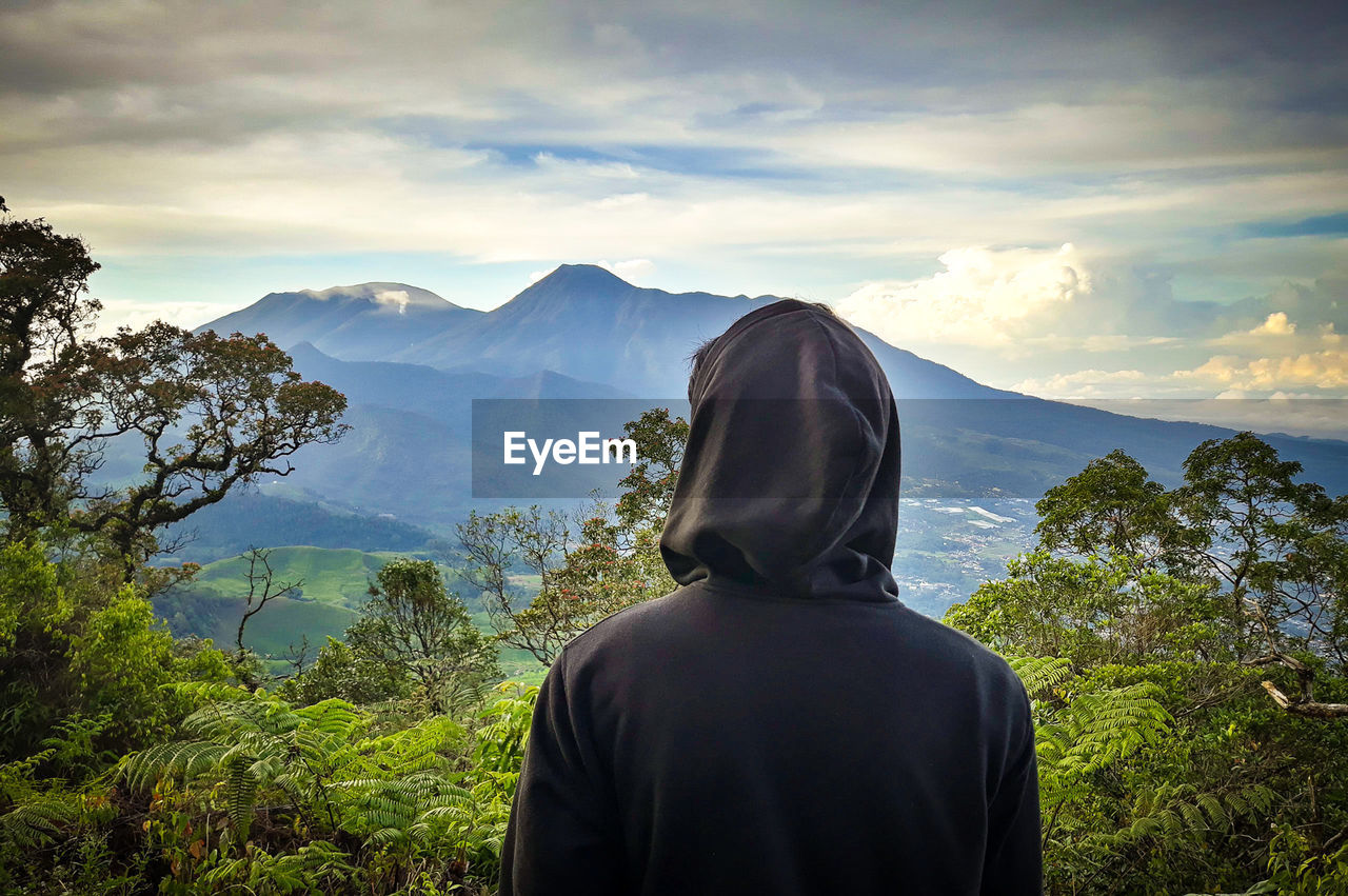 REAR VIEW OF MAN LOOKING AT MOUNTAIN RANGE AGAINST SKY