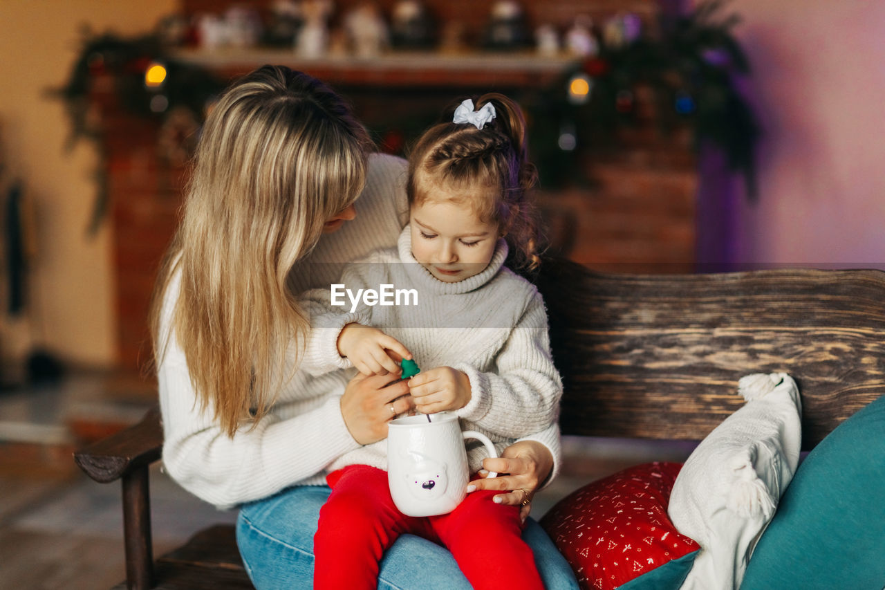 Portrait of a happy little girl sitting on her mother's lap and drinking hot chocolate on christmas