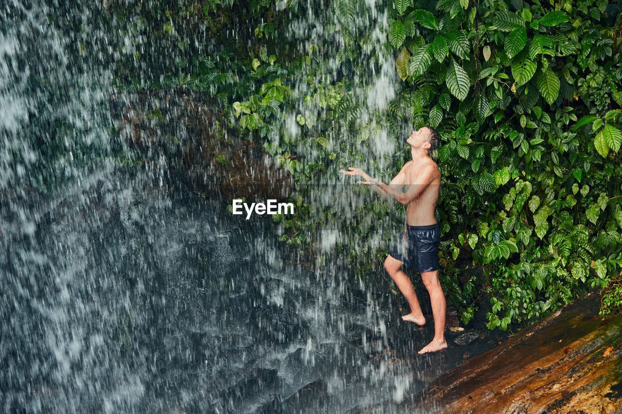 Full length of shirtless man standing under waterfall in forest