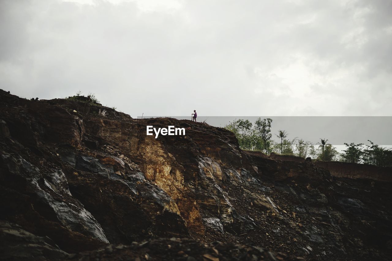 LOW ANGLE VIEW OF PERSON ON ROCK BY MOUNTAIN AGAINST SKY