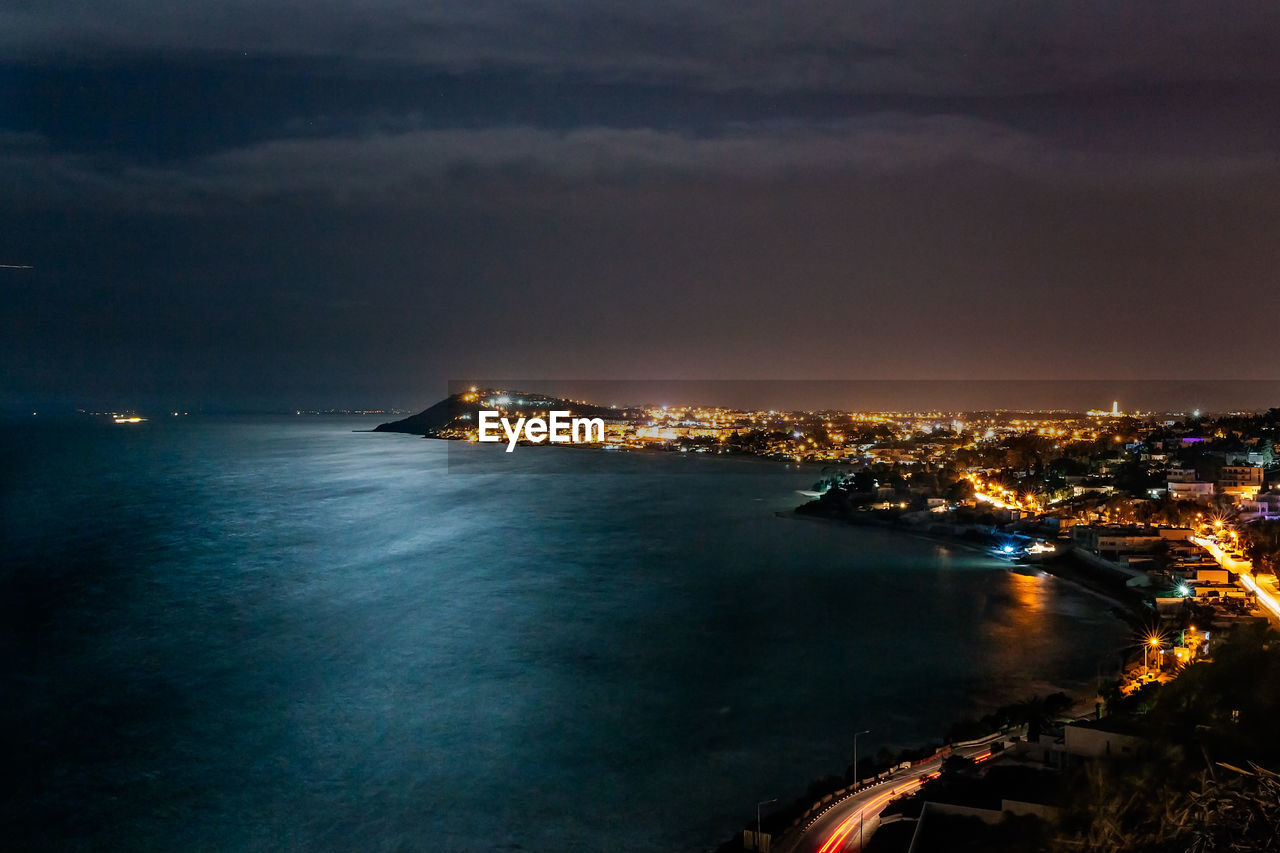 Illuminated city by sea against sky at night