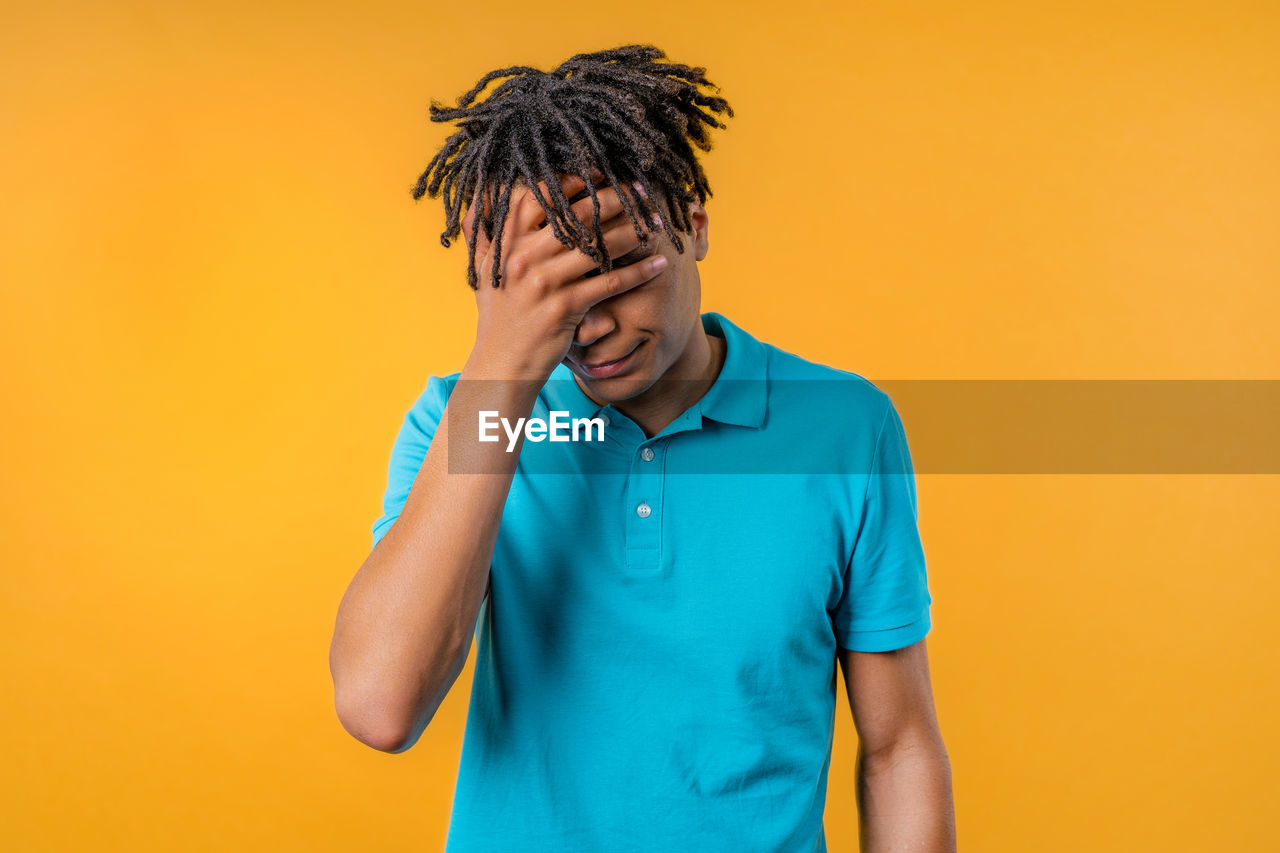 colored background, studio shot, one person, yellow, adult, emotion, young adult, indoors, men, negative emotion, frustration, waist up, emotional stress, sadness, portrait, yellow background, casual clothing, copy space, photo shoot, clothing, front view, depression - sadness, orange color, hand, person, head in hands, eyes closed, t-shirt, hairstyle, headache, facial expression, blue, worried, distraught, polo shirt, tired, standing, exhaustion