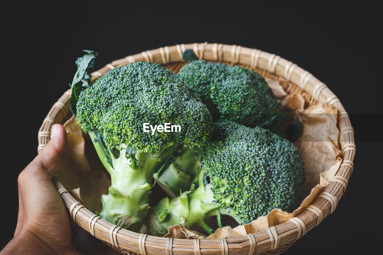 Cropped hand holding broccolis in basket against black background