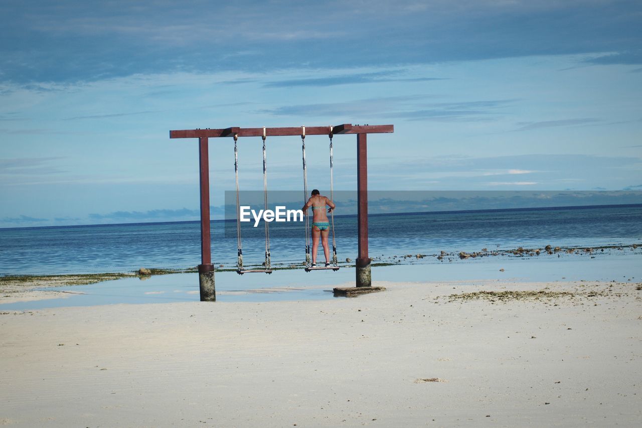 Rear view of woman in bikini standing on swing at shore of beach