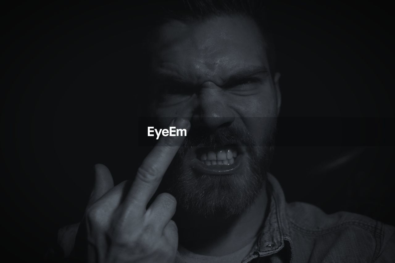 Close-up portrait of frustrated bearded man showing middle finger against black background