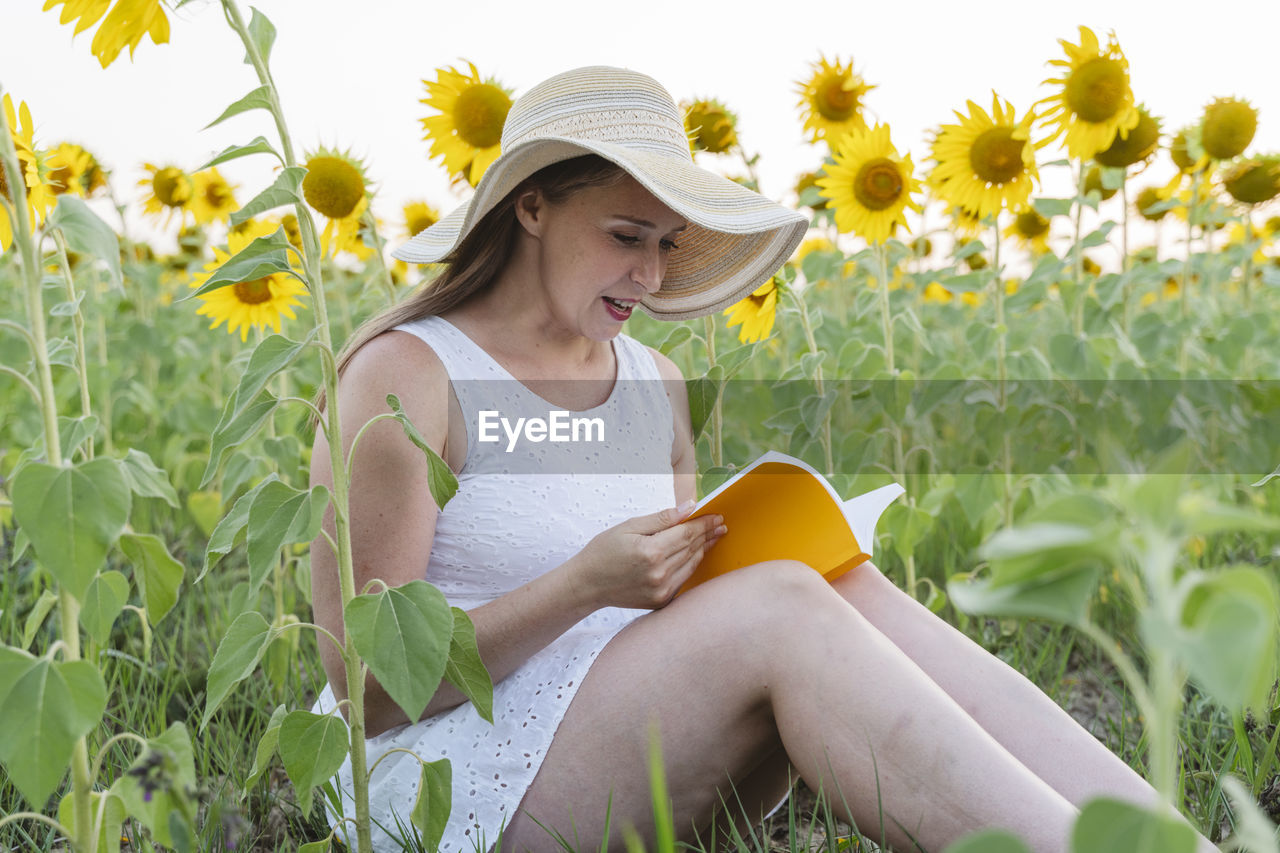 Woman with hat reading book sitting at sunflower field