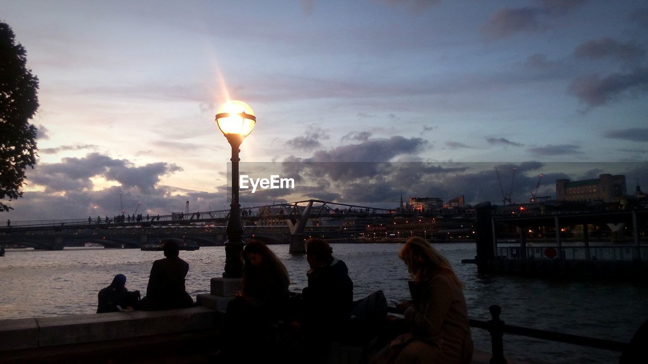 PEOPLE SITTING BY RIVER IN CITY AGAINST SKY