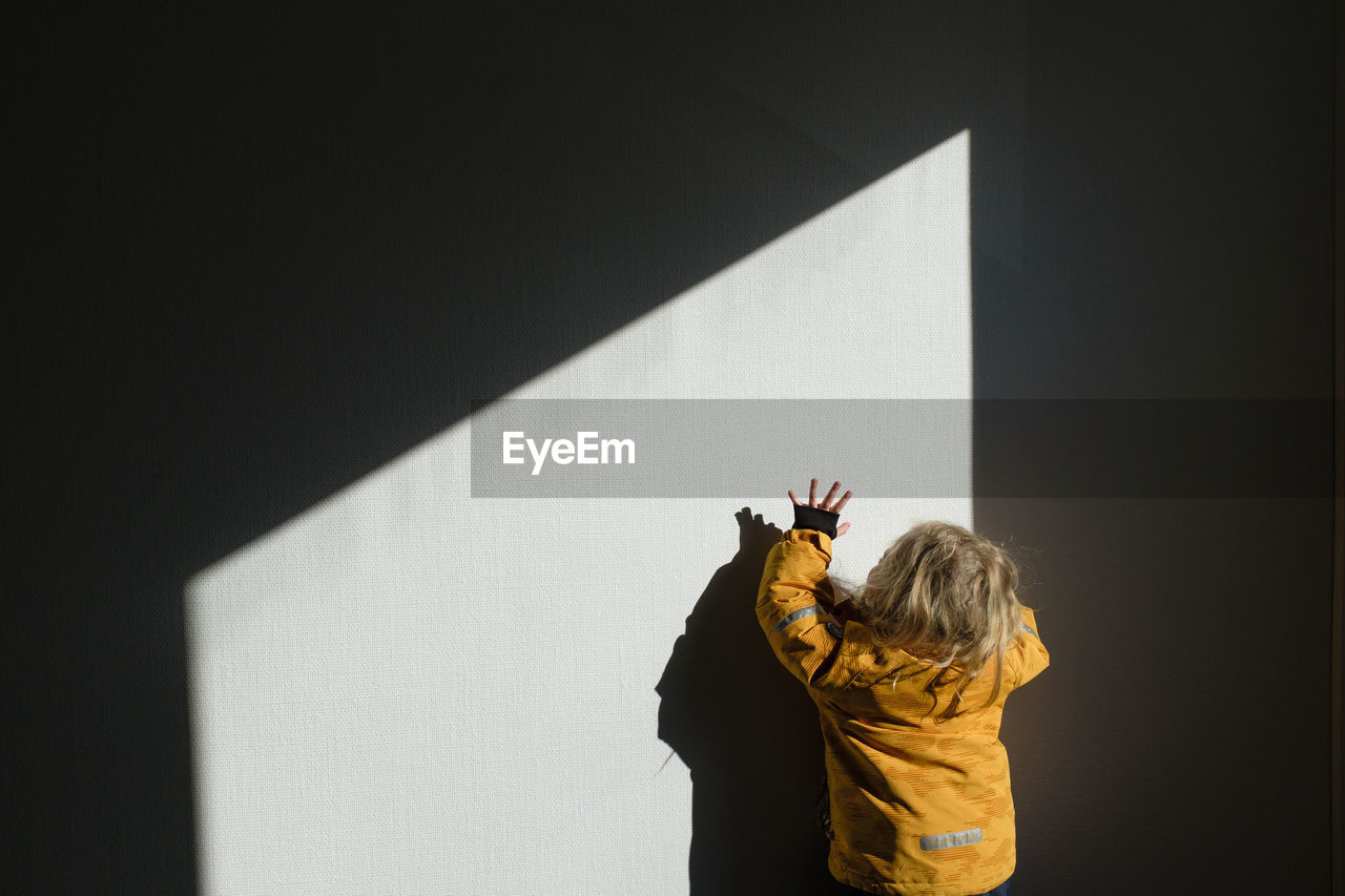 REAR VIEW OF WOMAN STANDING BY WALL WITH SHADOW